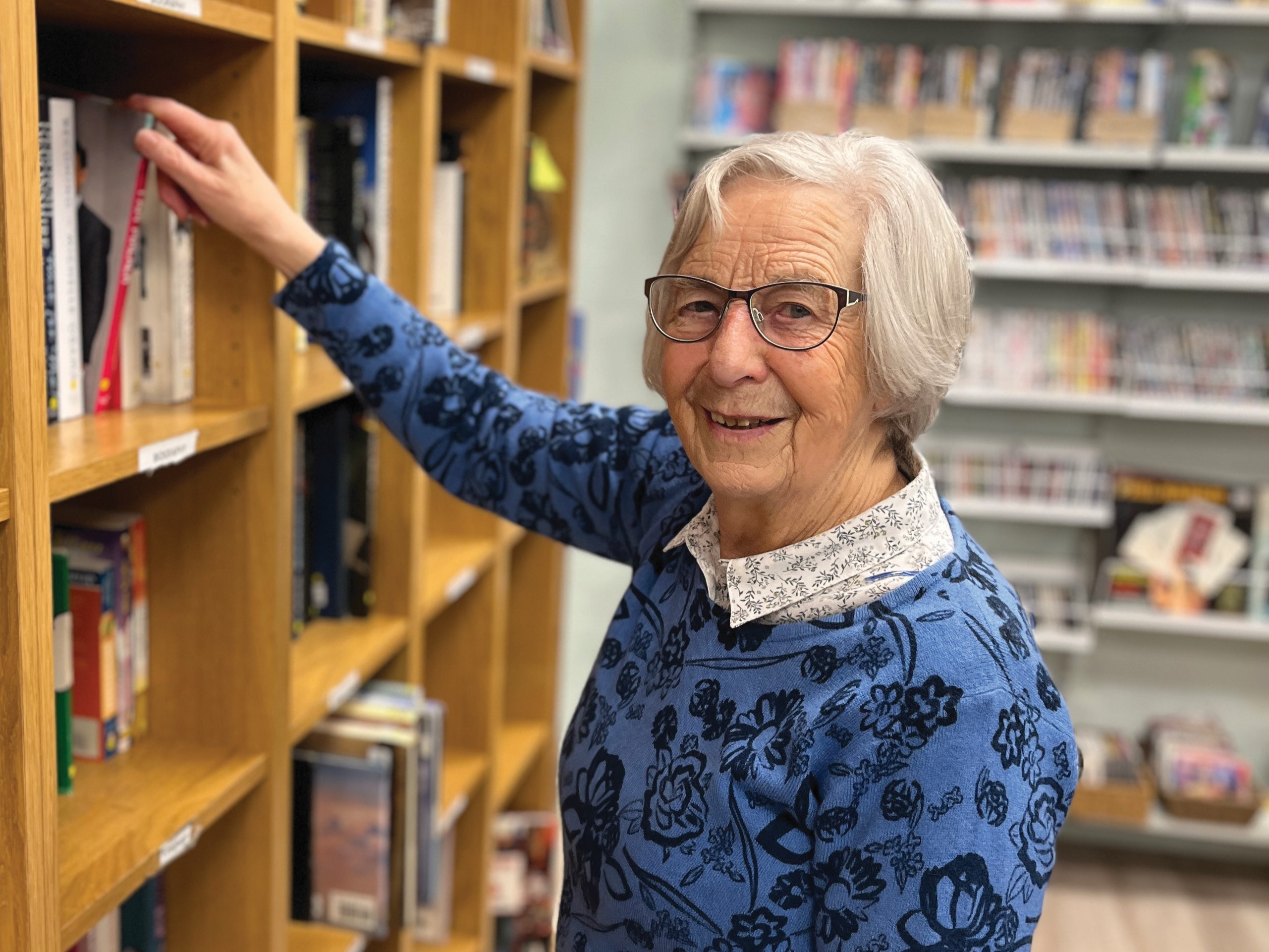 Shop volunteer in blue jumper placing a book on shelf in non-fiction section.