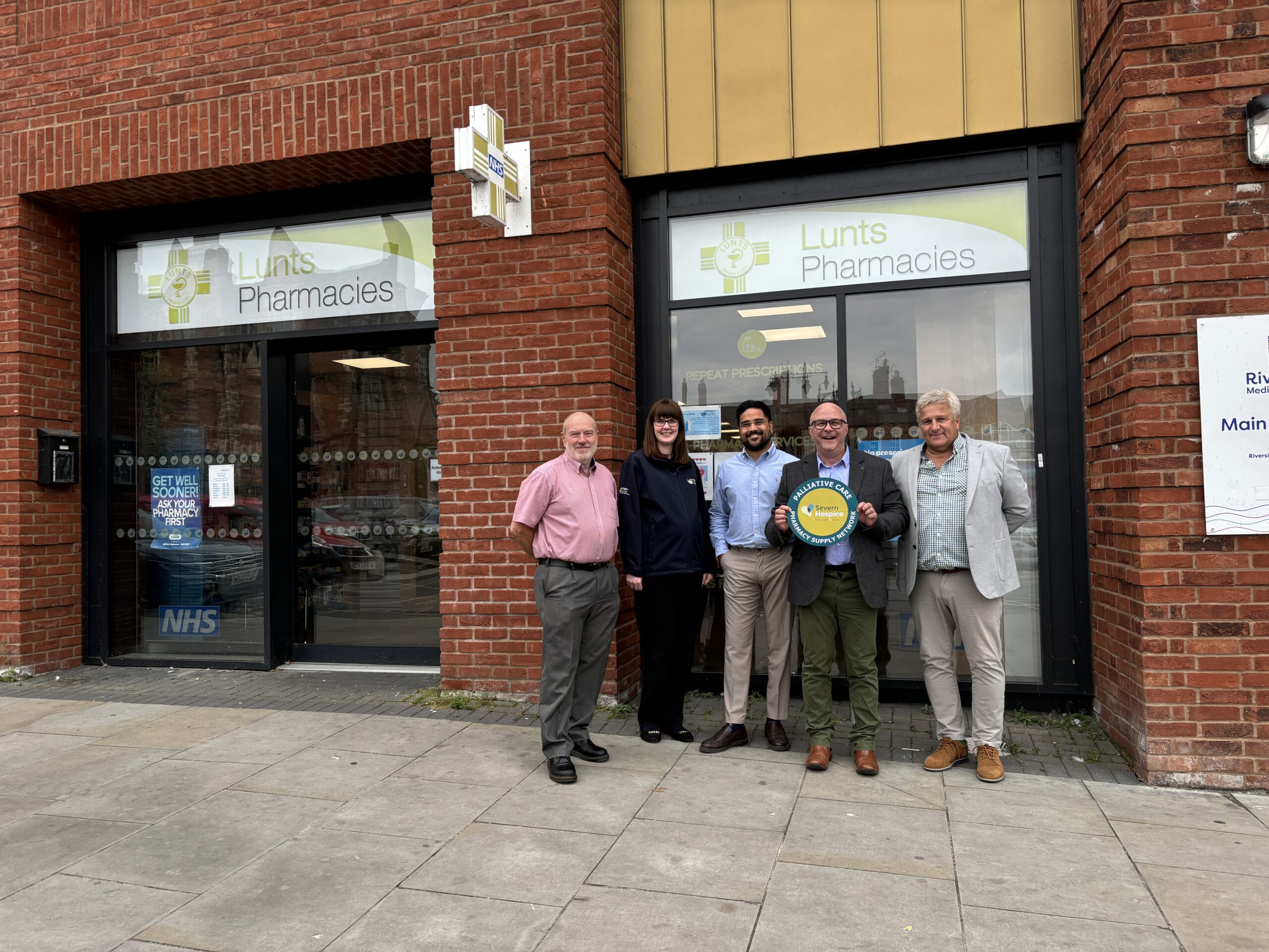 A group of people stand in front of a pharmacy, holding a window sticker.